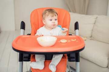 Cheerful happy child eats food by himself with a spoon. Portrait of a happy boy in a high chair for feeding.