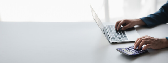 Businessman using a calculator to calculate numbers on a company's financial documents, he is...