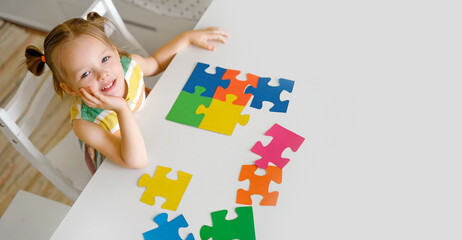 A cute beautiful little innocent girl is sitting at a table with large colorful puzzles. A puzzle for preschool development. The symbol of autism