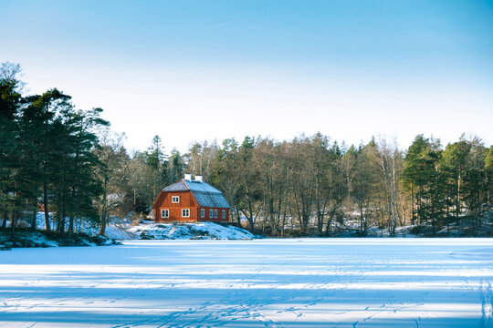 Traditional idyllic Swedish red wooden timber cabin house paint with falu red paint in winter landscape by frozen lake