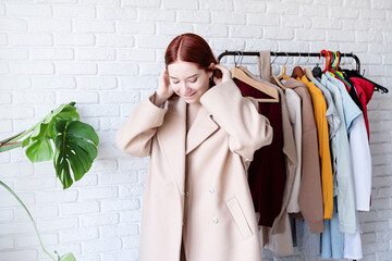 young beautiful woman wearing beige pastel coat, standing near clothes rack. Wardrobe change
