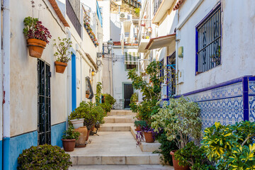 Fototapeta na wymiar Schmale Gasse durch spanische Altstadt, mit bunten Häusern und Pflanzen vor der Tür und auf dem Weg, bei sonnigem Wetter