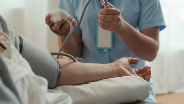 Close-up of young nurse in uniform pressing small rubber pump of tonometer while measuring blood pressure of mature sick female patient sitting in front of her during home visit