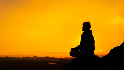 Silhouette of a person in the lotus position with orange sky and mountains in the background. Concept meditating at sunrise. Copy space on the right