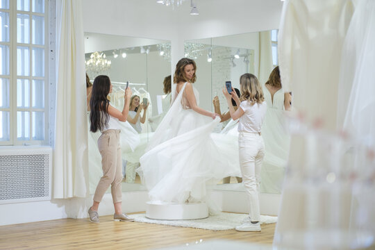 Women taking photographs of a female friend trying on wedding dress. Women in wedding dress fitting room.