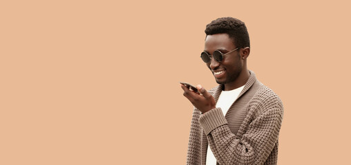Portrait of smiling young african man holding smartphone using voice command recorder, assistant or takes calling, looking at smartphone isolated on brown background