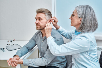 Experienced female doctor fitting Intra-The-Ear hearing aid into patient's ear while consultation in audiology clinic. ITE and ITC hearing aids - 568459742