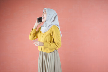 Obraz premium Beautiful Asian woman in yellow shirt and hijab smilling cheerfully holding smartphone on brown background 