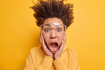 Crazy emotional young woman with curly hair standing up keeps hands on cheeks exclaims loudly keeps mouth widely opened wears transparent eyeglasses and hoodie isolated over yellow background