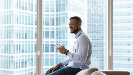 Happy millennial African man using mobile phone in at home, in hotel room, leaning on sofa, looking away, thinking on good news, posing against city building window view, enjoying communication