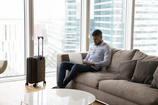 Focused Young Professional Man Working On Laptop On Business Trip, Using Computer In Hotel Room, Sitting On Sofa At Suitcase With Big Window, City View In Background