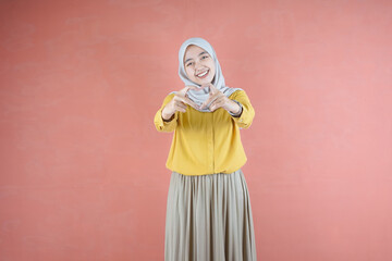 Beautiful Asian woman in yellow shirt and hijab feeling happy and romantic forming heart gesture expressing tender feelings on brown background 