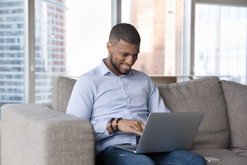 Cheerful busy African freelance entrepreneur man working from home, using laptop on sofa, typing,...