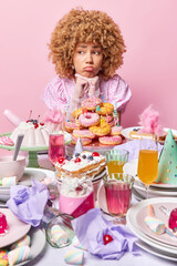 Upset curly haired young woman keeps hands under chin looks sadly aside feels lonely during birthday party poses near table full of desserts isolated over pink background. Celebration concept