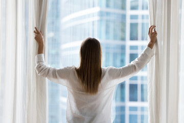 Rear view of young woman with long hair in casual opening drapes, parting white transparent curtains, veil at large window, enjoying view of urban buildings, city glass skyscrapers