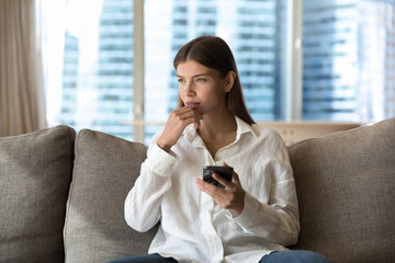 Pensive young millennial woman lost in thoughts holding cellphone, looking away, thinking, dreaming, touching chin, making decision, typing, making phone call, sitting on home couch