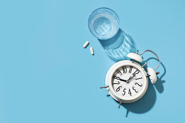 White alarm clock and pills and glass of water on blue background