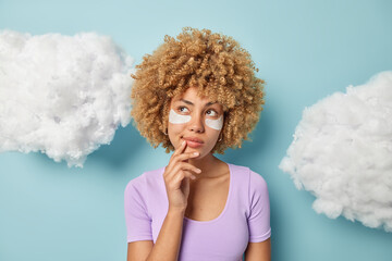 Thoughtful curly woman holds hand on chin considers something looks aside undergoes beauty procedures applies beauty patches dressed in purple t shirt isolated over blue background white clouds above