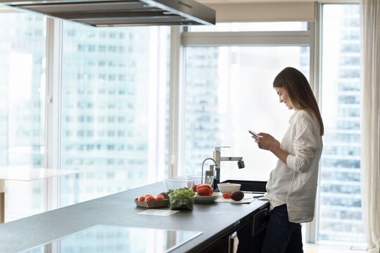 Happy young chef woman using mobile phone while cooking salad, browsing internet, searching for salad recipe, standing in modern home kitchen against large window with city view. Side shot
