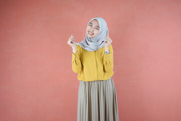 Fototapeta na wymiar Beautiful Asian woman in yellow shirt and hijab smiling cheerfully showing Korean heart with two fingers crossed, express joy and positivity over brown background 