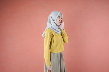 Fototapeta na wymiar Beautiful Asian woman in yellow shirt and hijab smiling cheerfully whispering over brown background