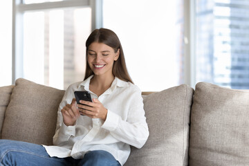 Cheerful beautiful millennial 20s girl typing on cellphone, sitting on home couch in city apartment, enjoying online Internet communication, touching screen, using financial ecommerce app