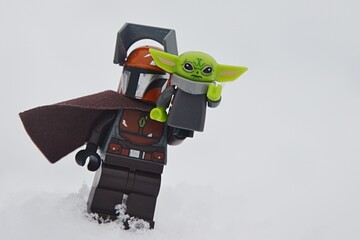 Naklejka premium LEGO Star Wars figure of Mandalorian in brown armor with cloak standing on deep snow with Baby Yoda aka The Child or Grogu on his right arm. 
