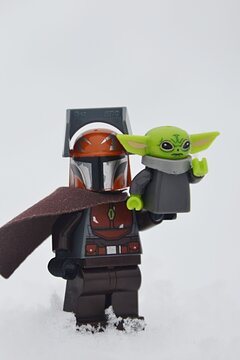Closeup view LEGO Star Wars figure of Mandalorian in brown armor with cloak standing on deep snow with Baby Yoda aka The Child or Grogu on his right arm. 