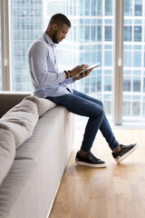 Focused young entrepreneur man using tablet at home for Internet communication, working on job tasks, making payment with bank application, sitting on sofa against big window city view