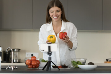 Cheerful healthy lifestyle blogger shooting video recipe, holding raw peppers, cooking salad from fresh vegetables, using organic food ingredients, looking, speaking at smartphone camera