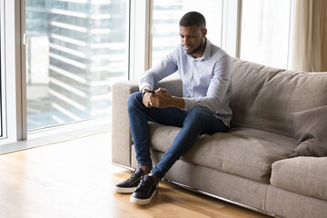 Handsome millennial young African freelance man typing on smartphone, sitting on home couch, using application on gadget, working on business tasks from urban city apartment
