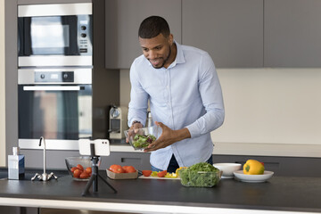 Handsome young Black cock blogger man recording healthy eating masterclass, video recipe of salad, using smartphone fixed on tripod, speaking, smiling, showing bowl with salad at screen, webcam