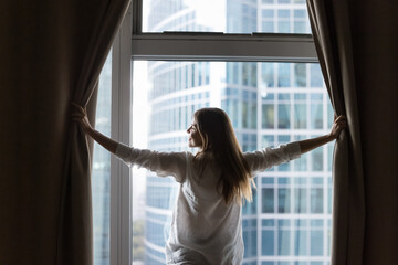 Happy pretty young woman standing at large window in new apartment, parting drapes at home, enjoying urban view from home in morning, breathing fresh air with open hands