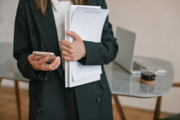 Young woman in jacket with documents using smartphone while standing at office desk.