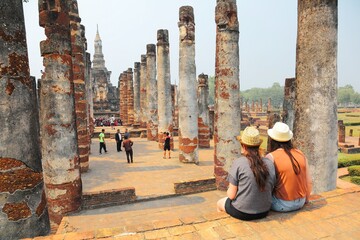Tourists sitting before ruined columns & enjoying the majestic view of Wat Mahathat, an ancient Buddhist temple in Sukhothai Historical Park, Thailand ~ A beautiful UNESCO heritage site in Thailand