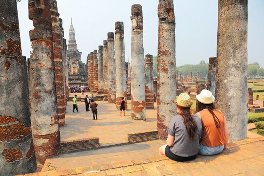 Tourists sitting before ruined columns & enjoying the majestic view of Wat Mahathat, an ancient Buddhist temple in Sukhothai Historical Park, Thailand ~ A beautiful UNESCO heritage site in Thailand