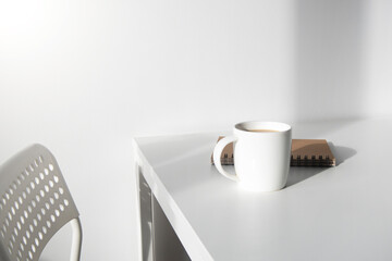 White cup of coffee, a notebook on a white table, there is a chair next to it.