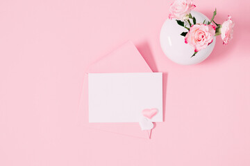 Top view on open pink envelope with paper card, bouquet of pink rose  flowers in vase and heart confetti on pastel pink table background. Birthday, Wedding, Mother's Day, Valentine's day, Women's Day.