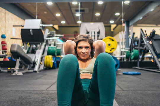Frontal image of a young female athlete at the gym sitting with her hands on the back of her neck while lifting her body to do sit-ups.