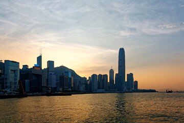 Sunset scenery of Hong Kong under dramatic sky, with famous landmark International Finance Center (IFC) standing among skyscrapers by Victoria Harbour & the setting sun with golden glow in background