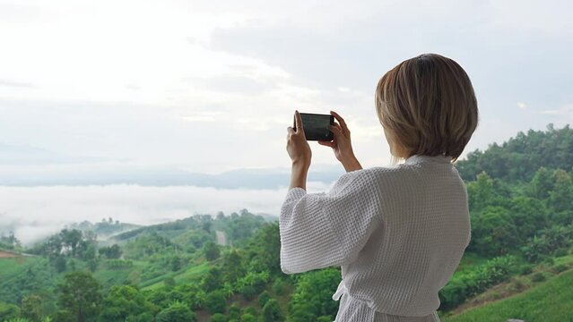 Oblique angle of the back of a young short-haired Asian woman in white bathrobe, standing outside hotel room on hills, using a smartphone slowly recording video of beautiful mountains with clouds.