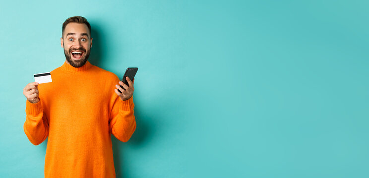 Online shopping. Surprised man holding mobile phone and credit card, paying in internet store, standing over light blue background