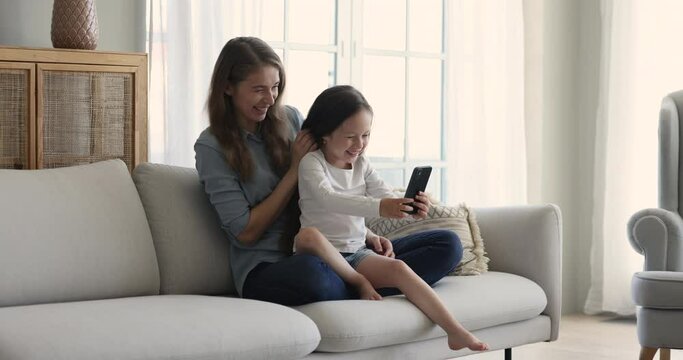 Happy woman her adorable daughter seated on mommys laps look at gadget screen, make selfie pictures on cellphone, rest on sofa, having fun using new amusing app spend leisure use modern wireless tech