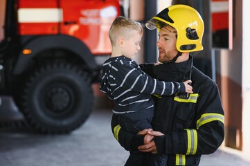 Obraz premium Portrait of rescued little boy with firefighter man standing near fire truck. Firefighter in fire fighting operation.
