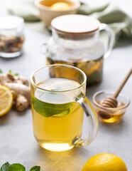 A glass of tea with fresh lemon, honey, mint and ginger on a light background with teapot and morning light close up.
