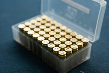 Selective focus and closeup of 45 rounds of 9 mm golden pistol ammo neatly placed vertically and showing the pistol primers in a transparent plastic ammunition box, on a blurred blue carpet floor.