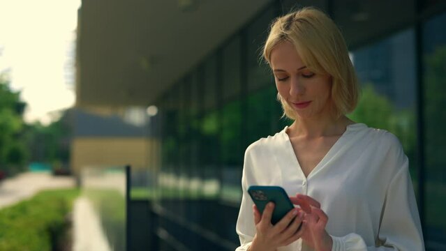 Business woman wearing formal suit standing near the office centre using the smartphone, texting with clients, swiping apps. People and technology concept.