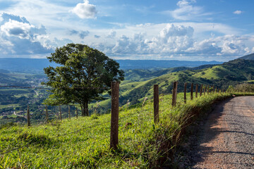 Fence and tree in the foreground with blue sky and hill in the background. Green mountains of Serra da Mantiqueira in the state of Minas Gerais, Brazil - obrazy, fototapety, plakaty