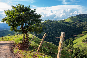 Fototapeta na wymiar Fence and tree in the foreground with blue sky and hill in the background. Green mountains of Serra da Mantiqueira in the state of Minas Gerais, Brazil