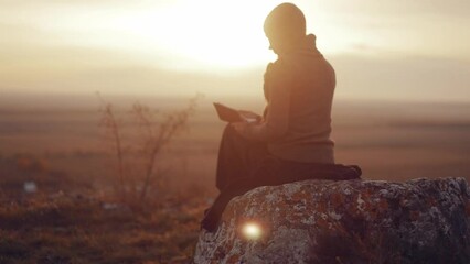 Young Christian woman girl reading the bible, praying, putting her hand on the bible, sitting on a rock in nature with an incredible view, sunset, peace and tranquility, faith. High quality 4k - Powered by Adobe